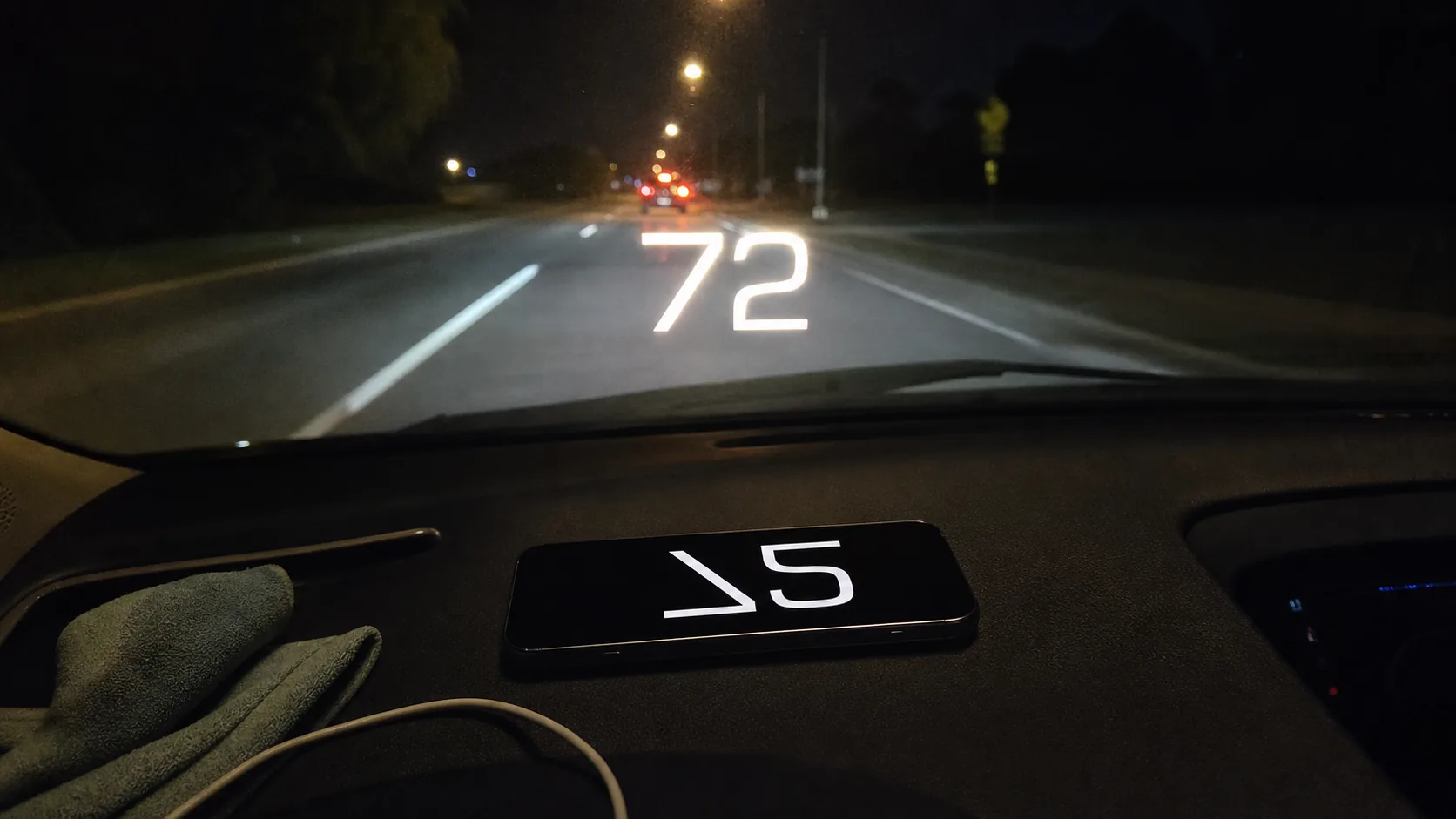 GPS Speedometer's HUD mode — iPhone face-up on a dashboard at night, the speed numeral mirrored onto the windshield above the steering wheel.